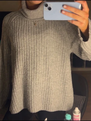 Grå ribbad polotröja - Mysig grå polotröja med ribbad struktur och lång ärm. Tröjan har en hög krage som ger extra värme och en loose passform som gör den skön att bära. Perfekt för kyliga dagar och enkel att matcha med jeans eller leggings.