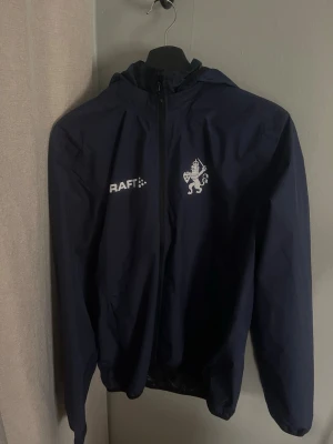 Mörk blå IFK Göteborg regnjacka från Craft - Säljer en mörkblå regnjacka från Craft i storlek M. Jackan har huva, hel dragkedja framtill och meshfoder på insidan för extra ventilation. Snygg IFK Göteborg logga på bröstet och diskreta detaljer. Perfekt för blåsiga dagar och sportiga aktiviteter. 