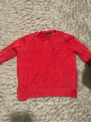 Röd kabelstickad tröja Polo Ralph Lauren - Kabelstickad röd tröja från Polo Ralph Lauren med klassisk marinblå logga broderad på bröstet. Tröjan har rund halsringning, ribbade muddar och lång ärm. Perfekt för dig som vill ha en tidlös och snygg look.