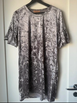 Grå/silvrig sammetklänning från DRAG - Supersnygg grå/silvrig kortklänning i sammet från DRAG. Klänningen har rund halsringning och korta ärmar med volangdetalj. Den har en glansig, krossad sammetslook som ger en cool och trendig vibe. Perfekt för dig som vill sticka ut lite extra.