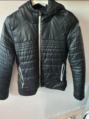 Svart quiltad pufferjacka från Adidas - Svart pufferjacka från Adidas med quiltade partier och huva. Jackan har vita dragkedjor framtill och på fickorna samt Adidas-logga på ärmen. Perfekt för kyliga dagar och har en sportig vibe.
