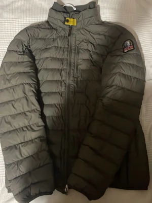 Grå dunjacka från Parajumpers - Säljer en grå dunjacka från Parajumpers med quiltad design och hög krage. Jackan har dragkedja framtill, en bröstficka med dragkedja och patch med logga på ärmen. Insidan är ljusgrå och har en säkerhetsficka. Perfekt för kalla dagar. (Jackan är i bra skick men på bild 5 ser man att att vänstra fickan är trasig. Det påverkar inte jackans funktion eller utseende i övrigt).