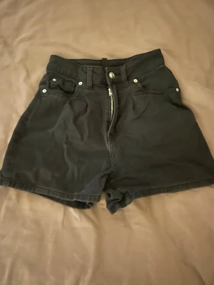 Svarta högmidjade jeansshorts - Säljer ett par svarta högmidjade jeansshorts i klassisk femficksmodell med knapp och dragkedja framtill. Shortsen har en loose passform och är tillverkade i bomull, perfekta för en chill och stilren look.