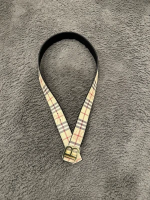 Rutig bälte med guldspänne - Snyggt bälte från Burberry med klassiskt rutigt mönster i beige, svart och rött. Bältet har ett stort guldigt B-spänne och är gjort i präglat skinn. Insidan är svart med diskret mönster. Perfekt accessoar för att lyfta din outfit.