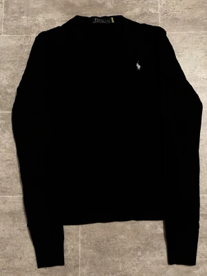 Svart kabelstickad tröja Polo Ralph Lauren - Svart kabelstickad tröja från Polo Ralph Lauren i storlek S. Klassisk modell med rund halsringning och broderad vit logga på bröstet. Tröjan är långärmad och har ribbade muddar vid ärmslut och nederkant. Perfekt för dig som gillar stilrena och tidlösa plagg. Passar M också.