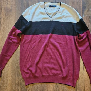 Randig v-ringad tröja Allen Solly XL - Snygg v-ringad tröja från Allen Solly i storlek XL. Tröjan har breda ränder i beige, svart och vinrött, med en liten broderad logga på bröstet. Långärmad och tillverkad i mjuk bomull, perfekt för dig som gillar stilrena färgkombinationer.