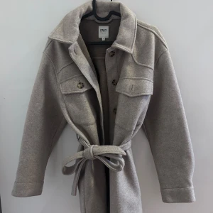 Beige kappa med bälte - Kappa i beige från ONLY med klassisk krage, stora bröstfickor, knappar framtill och ett matchande bälte i midjan. Jackan har lång ärm och är tillverkad i ett mjukt ullblandat material. Perfekt för lager-på-lager och snygg till jeans.