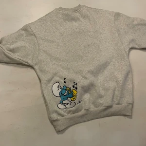 Grå Smurf & Butter sweatshirt S - Grå sweatshirt från Smurf &  Butter.. Stor tryckt illustration framtill samt mindre tryck på ryggen. Tröjan har rund hals, ribbade muddar och är långärmad. Oanvänd 