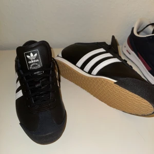 Adidas Italia svarta sneakers - Snygga Adidas Italia sneakers i svart skinn med klassiska vita ränder på sidorna och gummisula i brunt. Låg modell med snörning och retrovibbar, perfekt för dig som gillar stilrena och tidlösa sneakers. Adidas-logga på plösen och sidan.