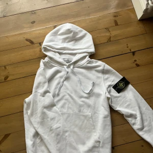 Stone Island hoodie  - Vit hoodie från Stone Island med klassisk patch på vänster ärm. Tröjan har huva med snörning och en stor magficka framtill. Mjuk bomullskvalitet och långärmad modell, perfekt för chill dagar. Snygg och enkel design som funkar till jeans eller joggers.