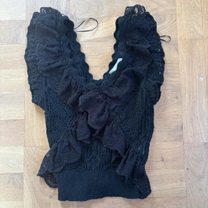 Svart spetsig blus med volanger🎀 - Säljer en svart blus med spets och stora volanger framtill. Toppen har v-ringad hals och är ärmlös med breda axelpartier. Detaljerad spets och ribbad nederkant ger en cool och romantisk vibe. Perfekt för dig som gillar statement-plagg!