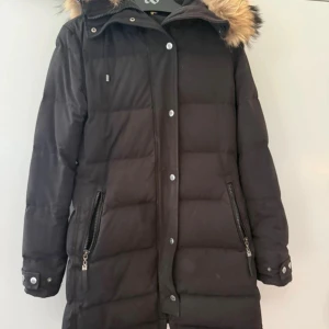 Svart dunjacka med fuskpäls från Hollies - Svart lång dunjacka från Hollies med stor huva och fuskpäls. Jackan har dragkedja och knappar framtill, två fickor med dragkedja och snygga detaljer vid ärmsluten. Perfekt för kalla vinterdagar och riktigt mysig att bära.