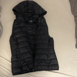 Svart dunväst från ONLY med huva - Svart quiltad dunväst från ONLY med huva och dragkedja framtill. Västen har två sidofickor och en sportig look med horisontella sömmar. Perfekt för lager-på-lager och chill dagar utomhus. Snygg och enkel att matcha med hoodies eller tröjor.