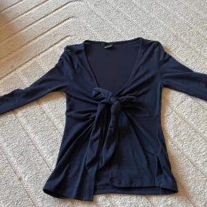 Svart omlottopp med knyt fram - Svart långärmad omlottopp med djup v-ringning och knyt framtill. Toppens material är mjukt och stretchigt, vilket gör den bekväm och smidig att bära. Perfekt för dig som gillar en stilren look med en twist.