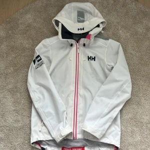 Vit regnjacka Helly Hansen  - Snygg vit regnjacka från Helly Hansen med huva och svart fleecefoder. Jackan har dragkedja med rosa detalj, logga på bröstet och ärmen samt justerbar huva. Jackan är i storlek s. Det finns några små fläckar på framsidan men den syns knappt