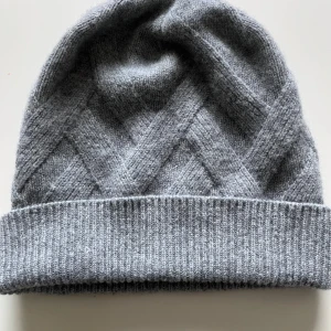 Grå Kashmir mössa med snygga mönster - Snygg grå mössa i kashmir med bred uppvikt kant och diskret kabelstickat mönster. Perfekt för kalla dagar och ger en stilren vibe till din outfit. Mjuk och varm känsla, passar både till jacka och hoodie.