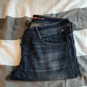 Mörkblå jeans från Jack & Jones - Snygga mörkblå jeans från Jack & Jones med klassisk femficksdesign och diskreta slitningar. Jeansen har raka ben och orangea sömmar vid linningen. Perfekta för en stilren och avslappnad look.