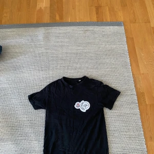 Svart t-shirt med Moncler-logga - Svart t-shirt från Moncler med korta ärmar och rund hals. På bröstet sitter en stor vit patch med Moncler-logga och coolt tryck. Perfekt för dig som gillar streetwear och vill ha något som sticker ut.