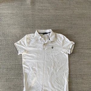 Vit Sail racing piké med svart detalj - Vit pikétröja med klassisk krage och svarta detaljer på ärmslut och krage. Tröjan har korta ärmar och två knappar framtill. Liten broderad logga på bröstet ger en clean look.