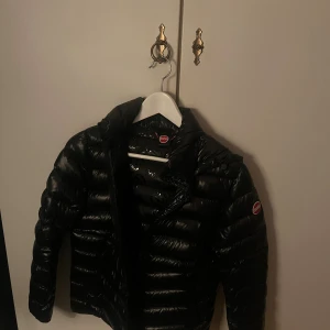 Svart pufferjacka från Colmar - Snygg svart pufferjacka från Colmar med glansig finish och klassisk logga på ärmen. Jackan har huva, dragkedja samt quiltad design. Perfekt för kalla dagar när du vill hålla stilen. Säljer då jag har köpt ny vinter jacka. Kan diskutera pris.💕