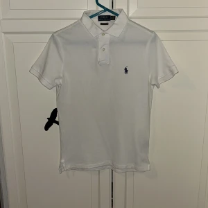 Vit pikétröja Polo Ralph Lauren XS - Klassisk vit pikétröja från Polo Ralph Lauren i modellen Custom Slim Fit. Tröjan har korta ärmar, krage med två knappar och den ikoniska broderade loggan i mörkblått på bröstet. Tillverkad i mjuk bomull, perfekt för en clean och stilren look.