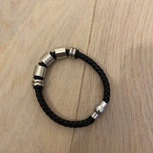Svart flätat armband med silverdetaljer - Snyggt svart armband med flätad design och coola silverfärgade metallringar och cylinderformade detaljer. Magnetlås i silver gör det enkelt att ta på och av. Perfekt accessoar för dig som gillar stilrena och moderna smycken.