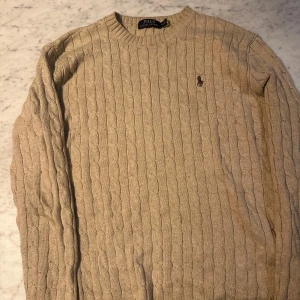 Beige kabelstickad tröja Polo Ralph Lauren - Kabelstickad beige tröja från Polo Ralph Lauren med klassisk rund halsringning och broderad logga på bröstet. Tröjan har lång ärm och är tillverkad i mjuk bomull, perfekt för lager-på-lager under höst och vinter.
