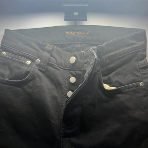 Svarta jeans från Nudie Jeans, - Svarta jeans från Nudie Jeans med klassisk femficksdesign och silverfärgade knappar. Jeansen är tillverkade av 100% ekologisk bomull och har Nudie Jeans logga på bakfickan. Passformen är normal och midjan är hög. Snygga kontrastsömmar och stilren look.
