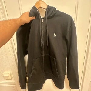 Svart hoodie från Polo Ralph Lauren(S) - Svart hoodie från Polo Ralph Lauren med klassisk vit logga på bröstet. Tröjan har dragkedja, huva med snörning och två stora fickor framtill. Tillverkad i mjuk bomull, perfekt för chill dagar. Snygg och enkel design som funkar till det mesta. Storlek S, skick bedöms enligt bilder