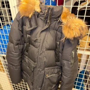 Svart dunjacka med pälsluva - Snygg svart dunjacka med stor fluffig päls runt luvan. Jackan har flera fickor med dragkedja och knappar, samt detaljer i svart skinn. Perfekt för kalla vinterdagar och har en riktigt maffig look.