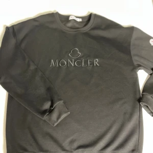 Svart sweatshirt från Moncler - Svart sweatshirt från Moncler med broderad logga på bröstet och klassisk Moncler-patch på ärmen. Tröjan har rund halsringning, ribbade muddar och är tillverkad i mjuk bomull. Perfekt för en clean och stilren look.