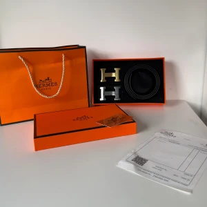 Svart H bälte med H-spänne - Exklusivt svart bälte från H i äkta skinn, levereras med två utbytbara H-spännen i guld och silver. Bältet har en stilren och klassisk design, perfekt för att lyfta din outfit med en lyxig touch. Kommer i originalförpackning med certifikat och kvitto.