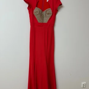 Röd långklänning med gulddetaljer - Snygg röd långklänning med korta ärmar och en unik guldfärgad paljettdekor framtill. Klänningen har en figurnära passform upptill och faller snyggt längs kroppen. Perfekt för dig som vill sticka ut med en elegant och glamorös look. Står att den är en storlek 38 men den är köpt från Istanbul och har turkiska mått, vilket motsvarar storlek 36 i eu storlek!
