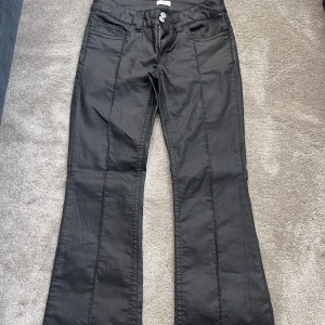 Svarta bootcut byxor från Nelly - Snygga svarta byxor från Nelly med bootcut-modell och markerade sömmar längs benen. Byxorna har två knappar framtill, dragkedja och klassiska fickor både fram och bak. Materialet har en lätt glansig finish som ger en cool look.