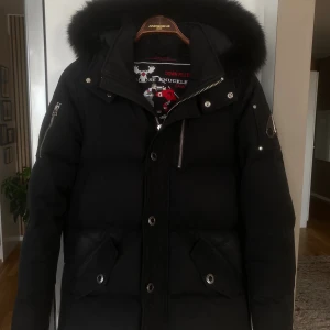 Moose Knuckles 3q jacka - Säljer en svart dunjacka från Moose Knuckles med fet pälskantad huva, stora fickor med tryckknappar och dragkedja. Jackan har snygga metalldetaljer på ärmen och broderad insida. Perfekt för kalla dagar och riktigt stilren look.