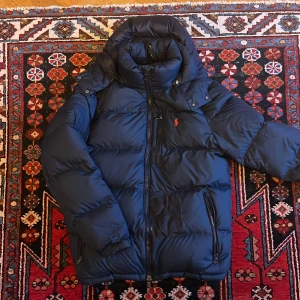 Mörkblå dunjacka från Polo Ralph Lauren - Säljer en mörkblå dunjacka från Polo Ralph Lauren med röd logga på bröstet. Jackan har hög krage, dragkedja framtill, tre fickor med dragkedja och en riktigt puffig look. Perfekt för kalla dagar och riktigt snygg till streetwear. Jackan är lagad med en svart lapp, syns på 3 bilden! Utöver lappen så är jackan i väldigt fint skick!