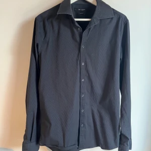 Svart skjorta Tiger of Sweden slim fit - Stilren svart skjorta från Tiger of Sweden i slim fit-modell. Skjortan har klassisk krage, knäppning framtill och långa ärmar. Materialet är mjukt och lätt, perfekt för en clean look.