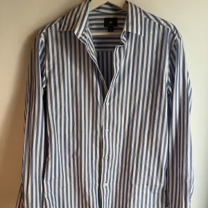 Blåvit randig skjorta från H&M - Snygg blåvit skjorta från H&M med klassiskt rakt snitt och långa ärmar. Skjortan har tydliga vertikala ränder, knappar framtill och en klassisk krage. Perfekt för dig som gillar stilrena och fräscha plagg.