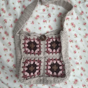 Beige och rosa virkad axelväska - Superfin handgjord axelväska i beige, rosa och bruna toner. Väskan är virkad med fyrkantiga mönster och har en bred axelrem samt en stor brun knapp som stängning. 🌸
