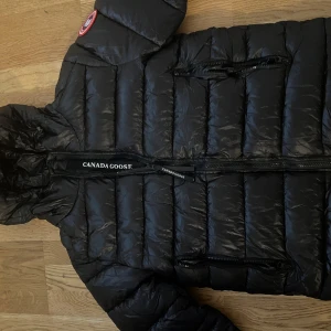 Svart dunjacka från Canada Goose - Säljer en svart dunjacka från Canada Goose med huva och flera dragkedjor framtill. Jackan har ett quiltat mönster och klassisk logga på axeln. Perfekt för kalla dagar med sin fluffiga och värmande design. Stilsäker look med tydliga Canada Goose-detaljer.