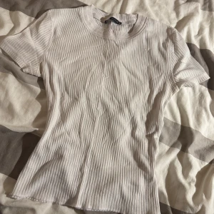 Vit ribbad topp från Zara - Säljer en vit ribbad topp från Zara med korta ärmar och rund halsringning. Toppen är i mjuk bomull och har en slim passform som sitter snyggt på kroppen. Perfekt att styla med jeans eller kjol för en clean look.