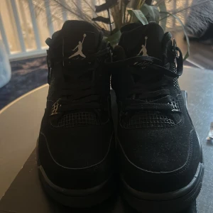 Svarta Nike Jordan 4 sneakers - Säljer ett par svarta Nike Jordan 4 sneakers med klassisk Jumpman-logga på plösen. Skorna har snörning, meshdetaljer och en robust platt sula. Materialet är en mix av mocka och syntet, vilket ger en cool och sportig vibe. Perfekta för dig som gillar streetwear och vill sticka ut.