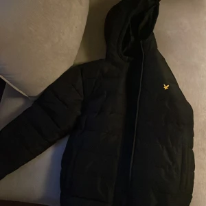 Svart pufferjacka från Lyle & Scott - Svart pufferjacka från Lyle & Scott med huva och gul fågel-logga på bröstet. Jackan har quiltad design, dragkedja framtill och långärmade ärmar. Perfekt för kalla dagar och har en clean, stilren look som funkar till streetwear.