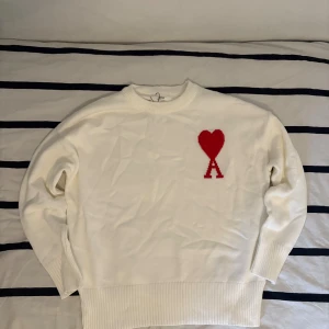 Vit stickad tröja från Ami Paris - Vit stickad tröja från Ami Paris med röd hjärtlogga och bokstaven A på bröstet. Tröjan har rund hals, ribbade muddar och är långärmad. Perfekt för dig som gillar stilrena plagg med en twist. Materialet är mjukt och känns lyxigt mot huden. Hör av dig om du har några frågor
