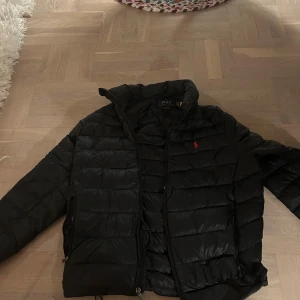 Svart pufferjacka från Polo Ralph Lauren - Svart pufferjacka från Polo Ralph Lauren med röd logga på bröstet. Jackan har dragkedja framtill, ståkrage och två sidofickor med dragkedja. Perfekt för kalla dagar och har en clean, sportig look som funkar till jeans och sneakers.