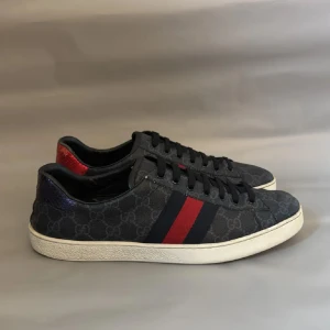 Gucci sneakers med GG-mönster - Snygga Gucci sneakers i mörkblått med GG-monogram över hela skon. Sidorna har klassiska röd-blå ränder och hälarna är i metallic röd och blå med Gucci-logga. Skorna har snörning, platt vit sula och rund tå. Materialet är canvas och skinn med reptilstruktur på hälen.