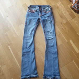 True Religion ljusblå bootcut jeans - Säljer ett par ljusblå jeans från True Religion med färgglada, grova sömmar i rosa och orange. Modellen är bootcut med låg midja och klassiska fickor med dekorativa knappar och broderade detaljer. Jeansen är i mjukt denim och har en riktigt unik look. Dom är i storlek 23. Har sprätta fållen för att dom var lite korta för dottern.