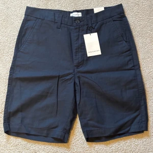 Marinblå shorts från These Glory Days - Snygga marinblå shorts från These Glory Days i storlek 48. Klassisk rak modell med midwaist, bälteshällor och knappstängning fram. Tillverkade i linneblandning av linne och bomull, med två sidofickor och två bakfickor. Perfekta för en chill sommardag.
