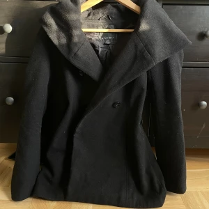 Svart kappa med stor huva från Amisu - Svart kappa från Amisu med stor, snygg huva och dubbelknäppning framtill. Jackan har en klassisk, stilren look och är tillverkad i ullblandning som håller dig varm. Perfekt för dig som gillar en tidlös och clean stil.