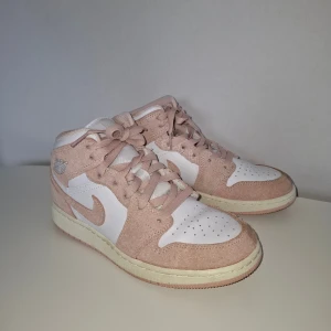 Nike Jordan 1 Mid rosa sneakers - Nike Jordan 1 Mid sneakers i ljusrosa och vitt med snygga mockadetaljer och klassisk snörning. Skorna har en rund tå, platt sula och ikonisk Jordan-logga på sidan. Perfekt för dig som vill ha en clean och trendig look med lite extra färg. Säljer dotterns skor som endast blev använd någon enstaka gång innan de blev för små. 
